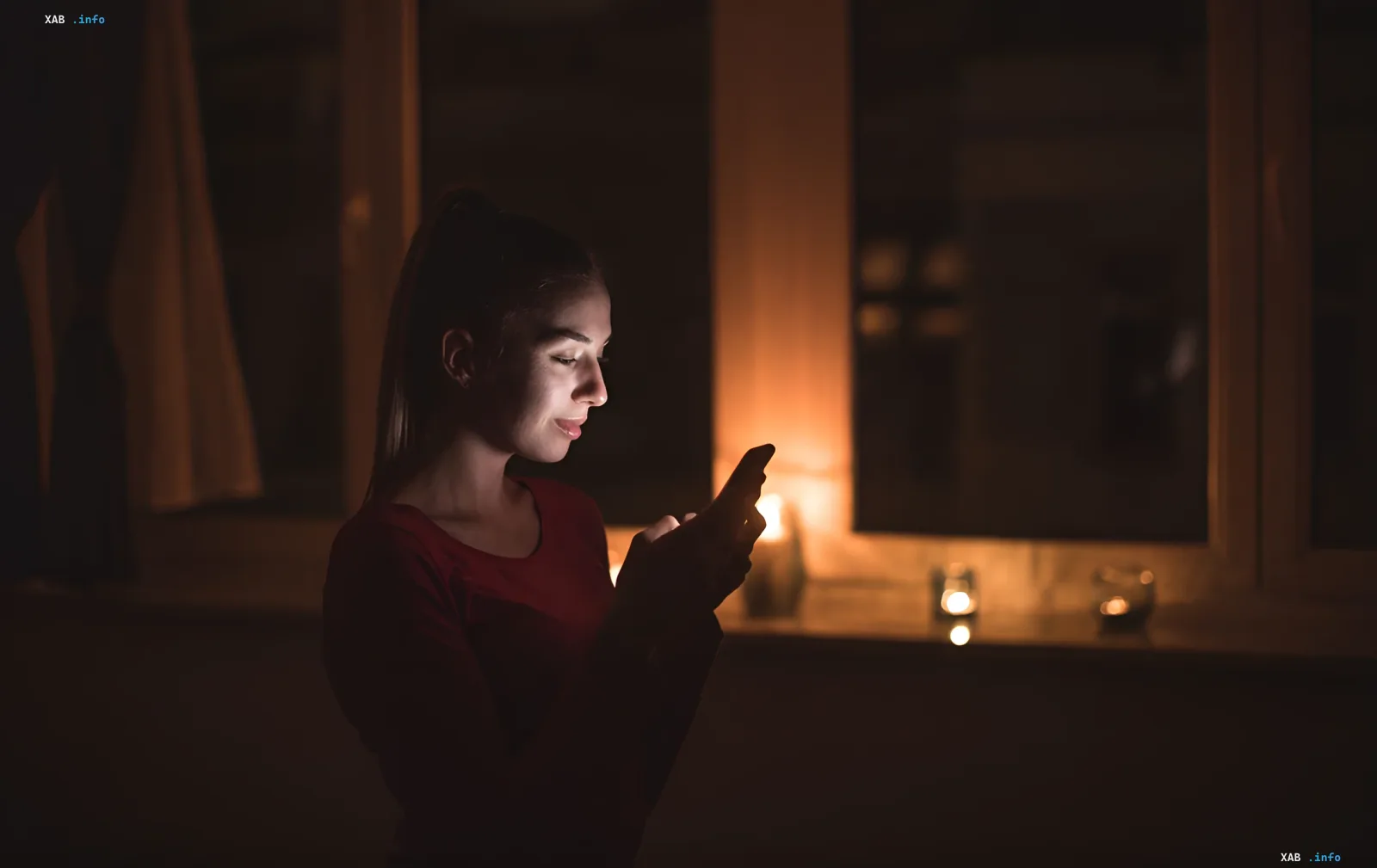 Женщина в темноте использует смартфон, освещенный экраном, на фоне горящих свечей, символизируя отключения электроэнергии летом.