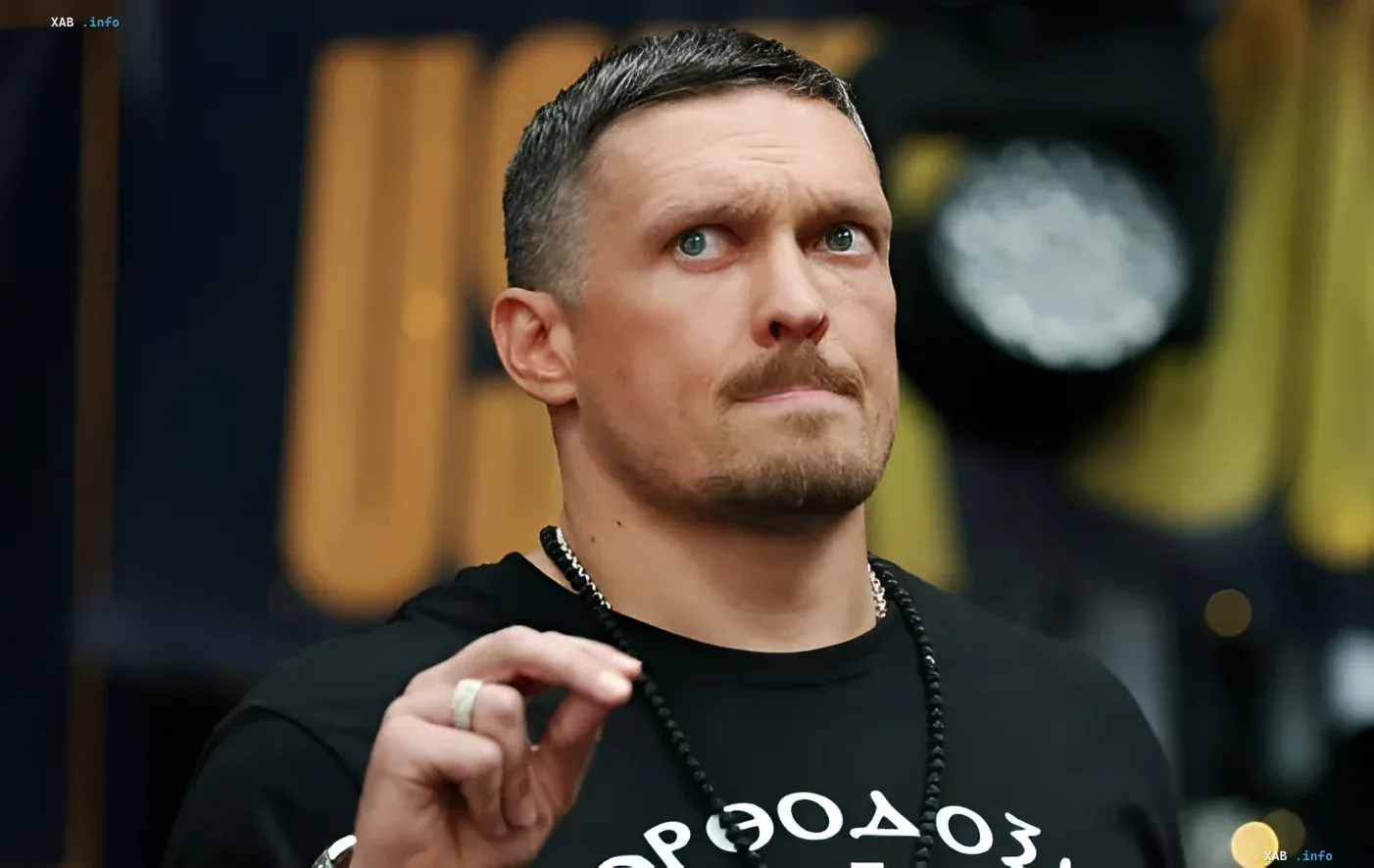 Александр Усик в черной футболке с надписью 'Орфей' на фоне пресс-конференции, задумчиво смотрит в сторону, держа бусы — анонс последнего боя