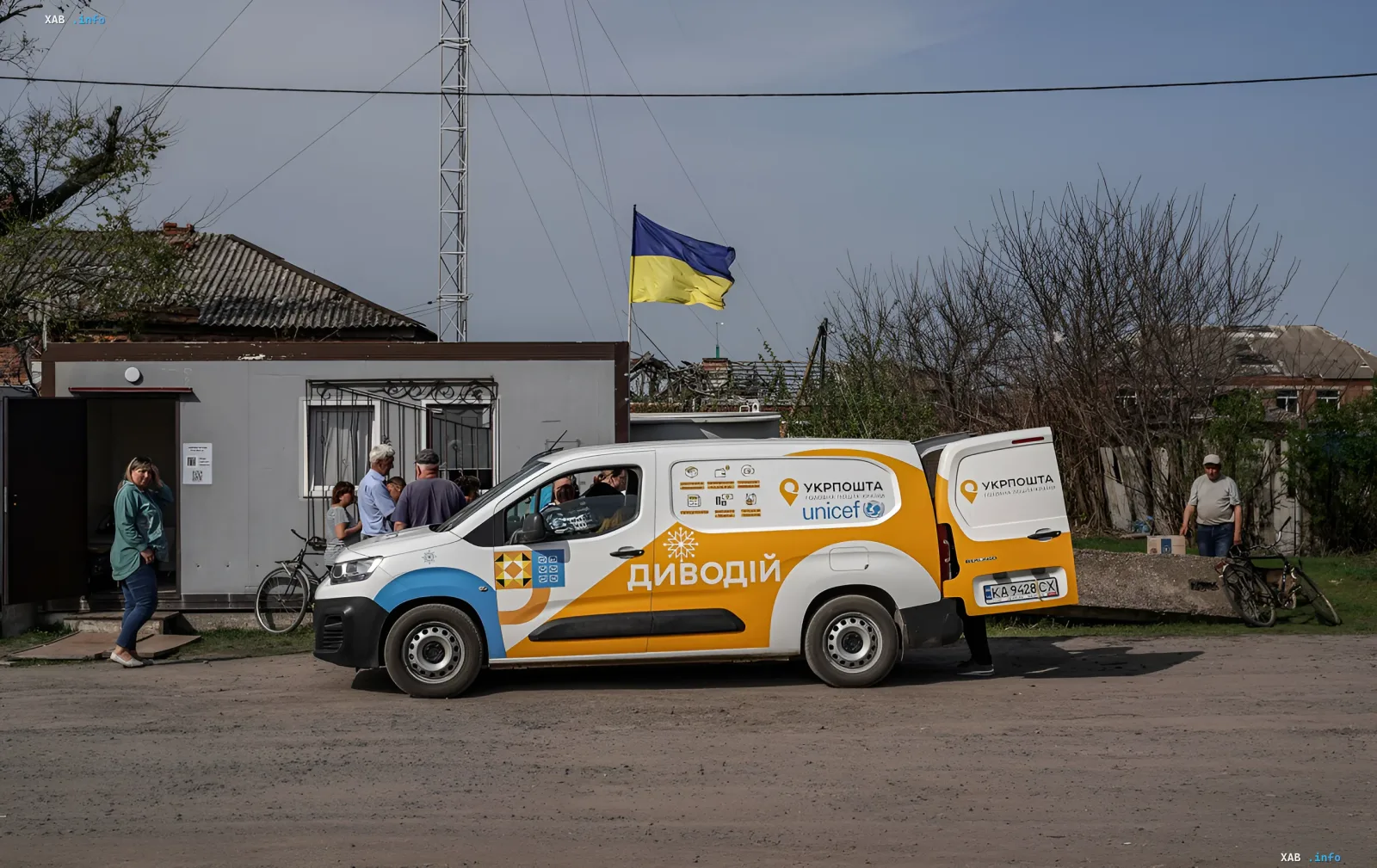 Грузовой фургон Укрпочты с логотипом UNICEF и надписью «Диводій» стоит у сельского почтового отделения, где люди получают посылки. На фоне украинский флаг и сельская местность.