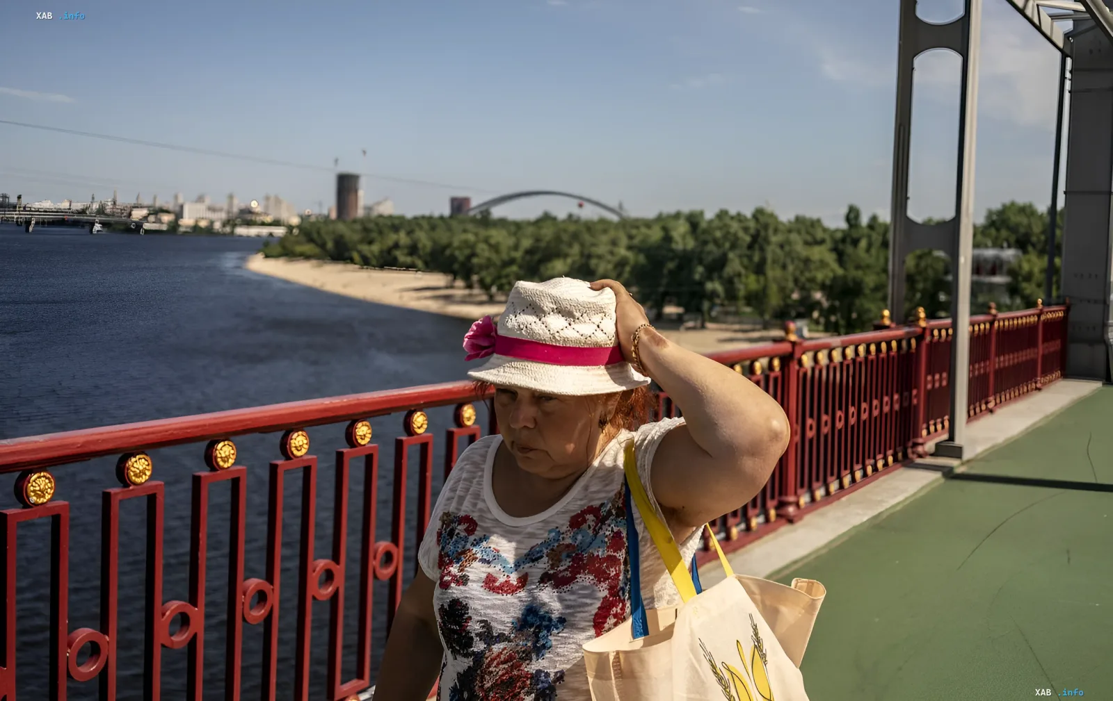 Женщина в соломенной шляпе с розовой лентой и цветной футболкой идет по мосту над рекой, прикрывая голову от палящего солнца — иллюстрация к новости о жаре в Украине +38°C