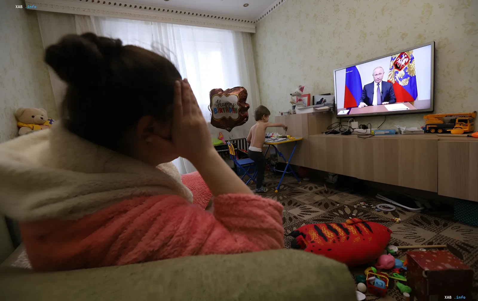 A woman watches anxiously as Vladimir Putin addresses the nation on TV, while a child plays in the background in a room with a 'Happy Birthday' balloon