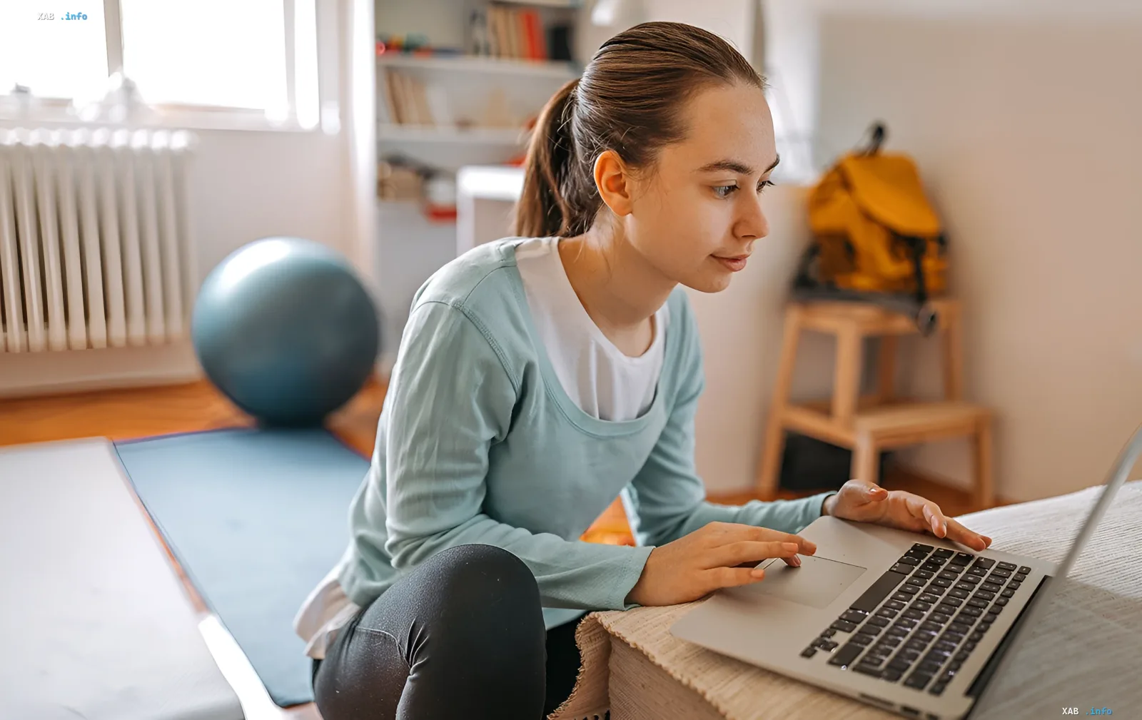Student using laptop in room with exercise ball and mat, choosing university by sports infrastructure via SportOsvita map