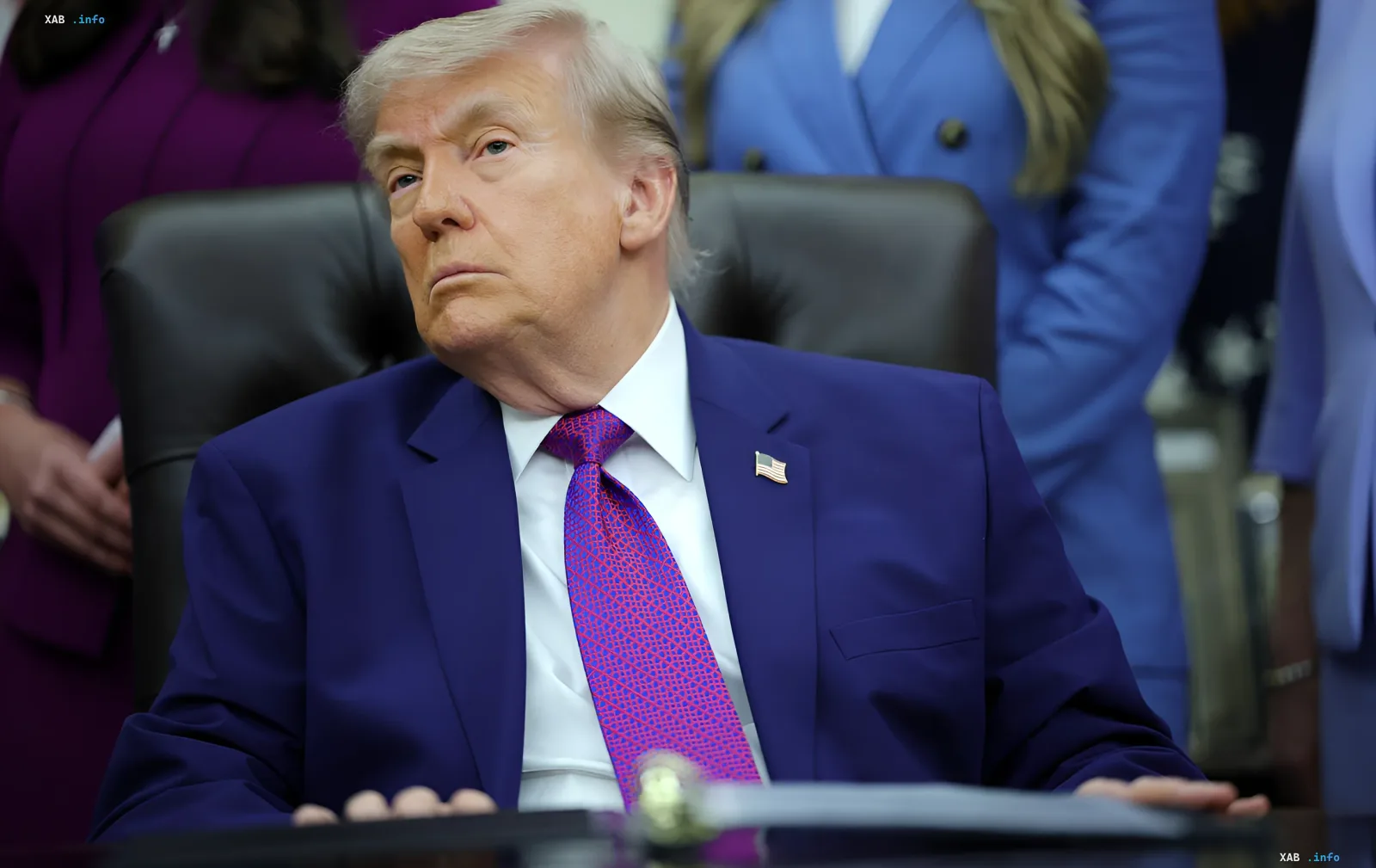 Дональд Трамп у синьому костюмі та червоному краватці сидить за столом, задумливо дивлячись убік, на фоні офіційного середовища з людьми в ділових костюмах.