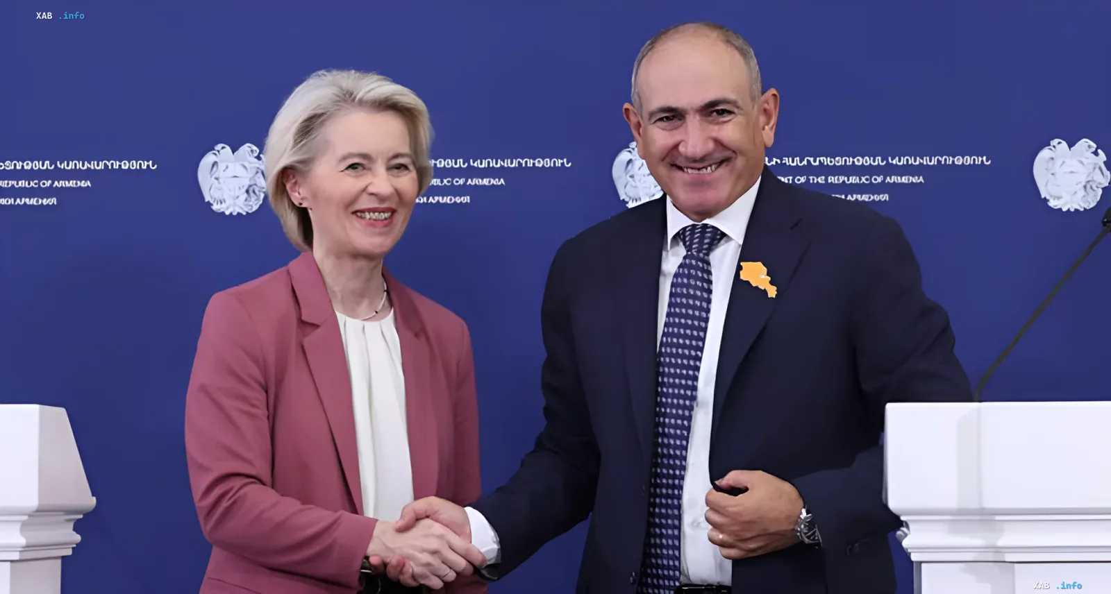 Голова Європейської комісії Урсула фон дер Ляйен та прем'єр-міністр Вірменії Нікол Пашинян тримаються за руки на фоні емблеми Республіки Вірменія, символізуючи поглиблення торгових відносин ЄС та Вірменії.