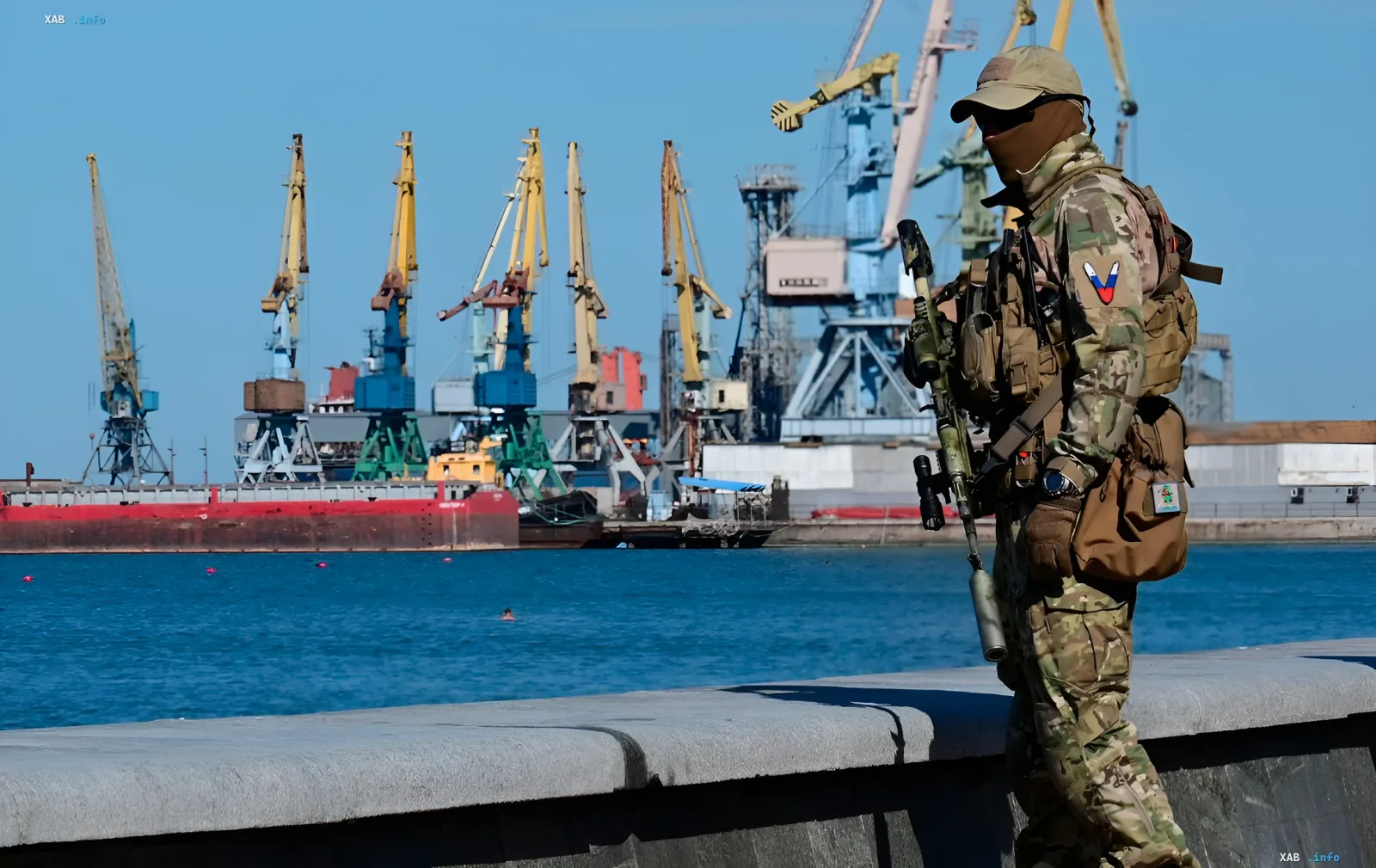 Военный в камуфляже с оружием стоит на набережной в Крыму, на фоне портовых кранов, готовясь к возможной высадке десанта ВСУ и атакам дронов.