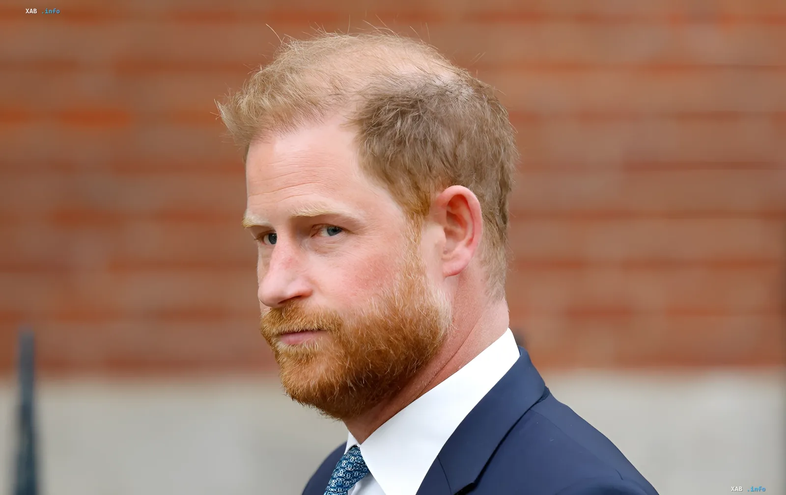 Prince Harry in a business suit against a brick wall, arriving in the UK without Meghan and children