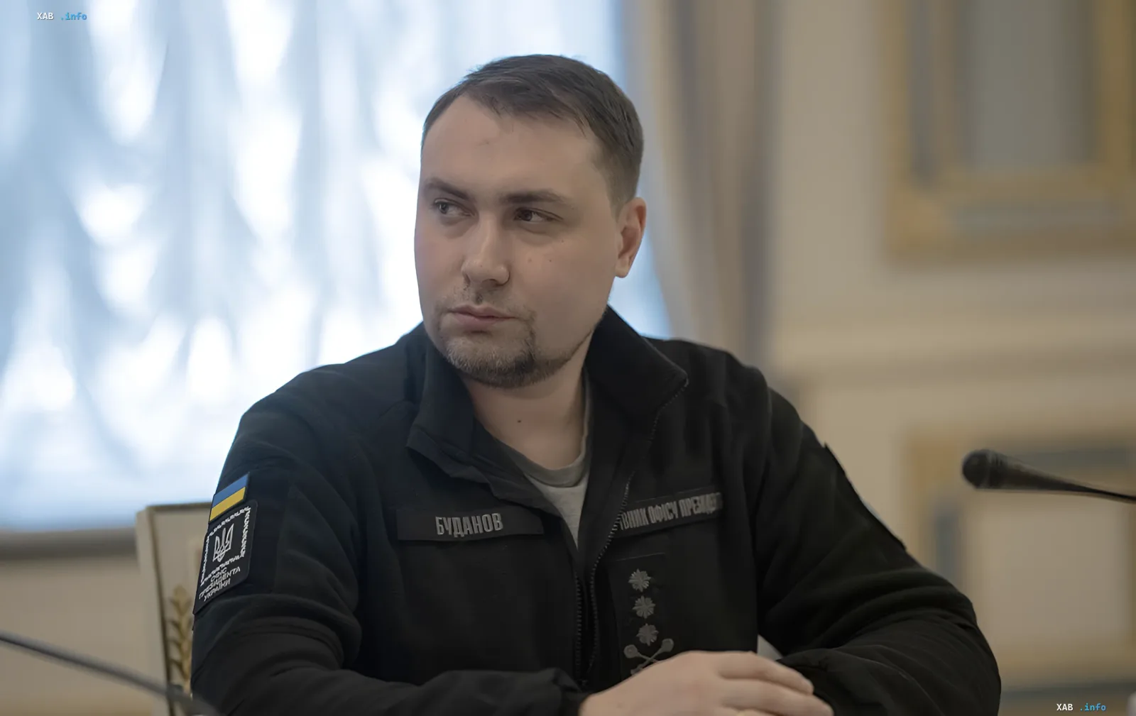 Kyrylo Budanov, head of Ukraine's Main Intelligence Directorate, in uniform with a 'Budanov' patch and Ukrainian emblem, against a backdrop of a microphone and formal setting, illustrating his role as a subordinate of President Zelenskyy.