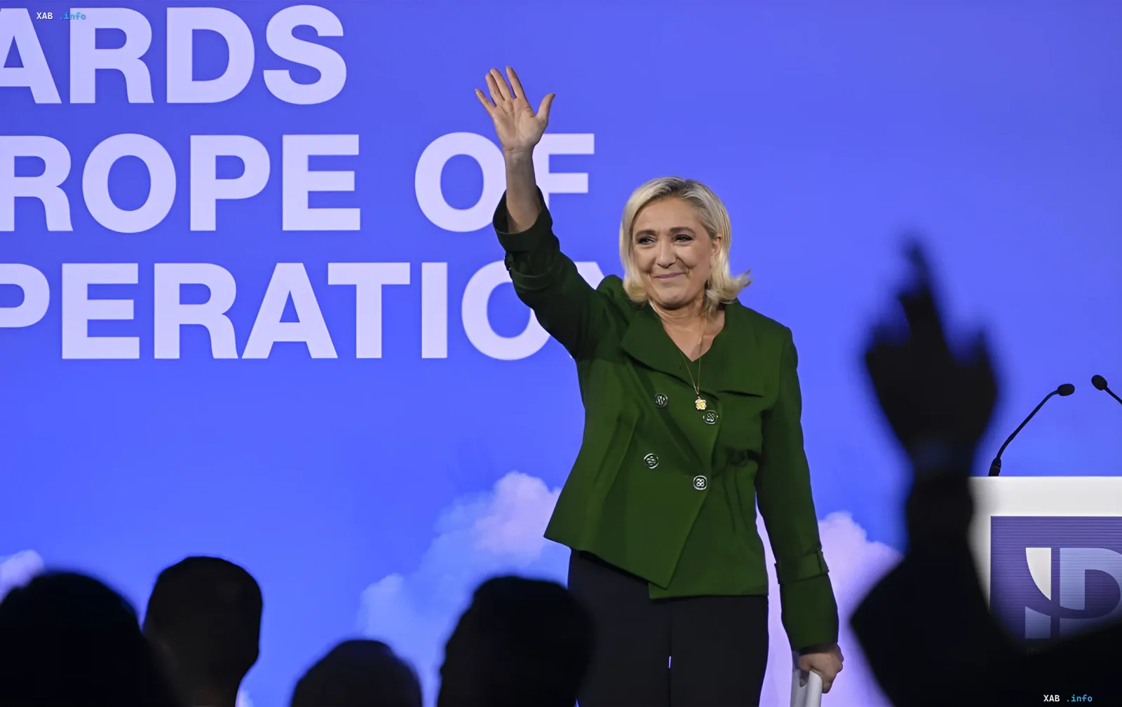 Marine Le Pen in a green jacket waves at an audience against a blue screen with the text 'AWARDS EUROPE OF COOPERATION', announcing her intention to run for President of France in 2027.
