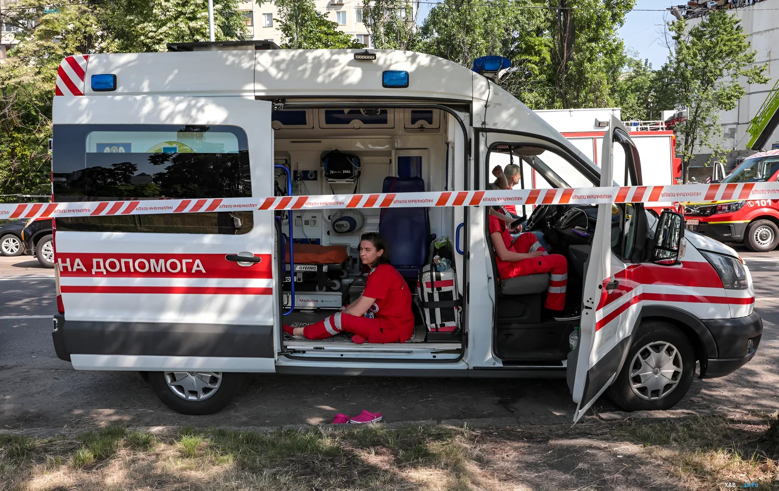 Медики в красной форме сидят в открытой машине скорой помощи после атаки дрона на маршрутку в центре Херсона, рядом — лента «НА ДОПОМОГА» и следы разрушений