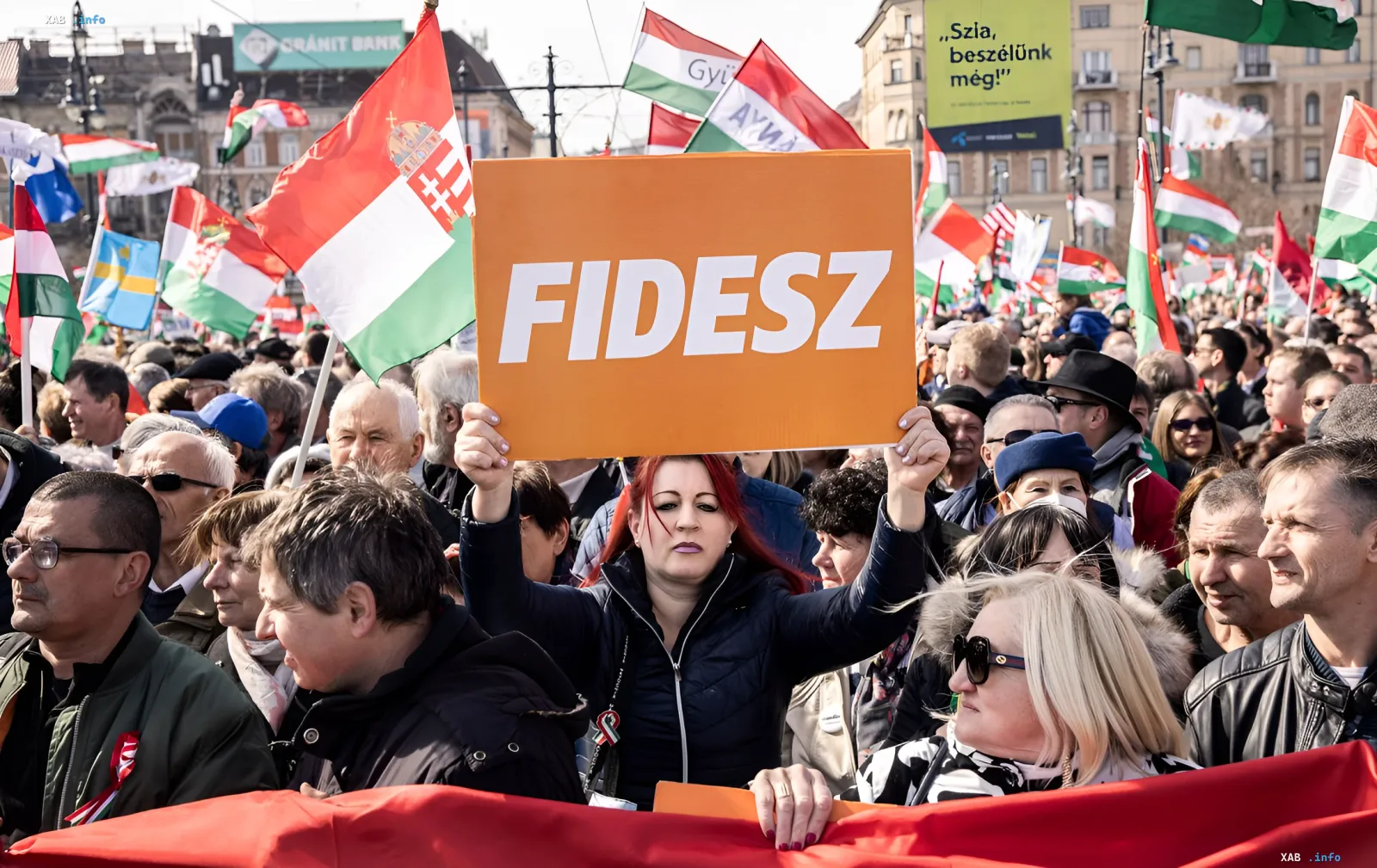 Supporters of Fidesz party at a rally in Hungary holding flags and an orange sign, symbolizing political backing of the regime linked to propaganda on state TV channel M1