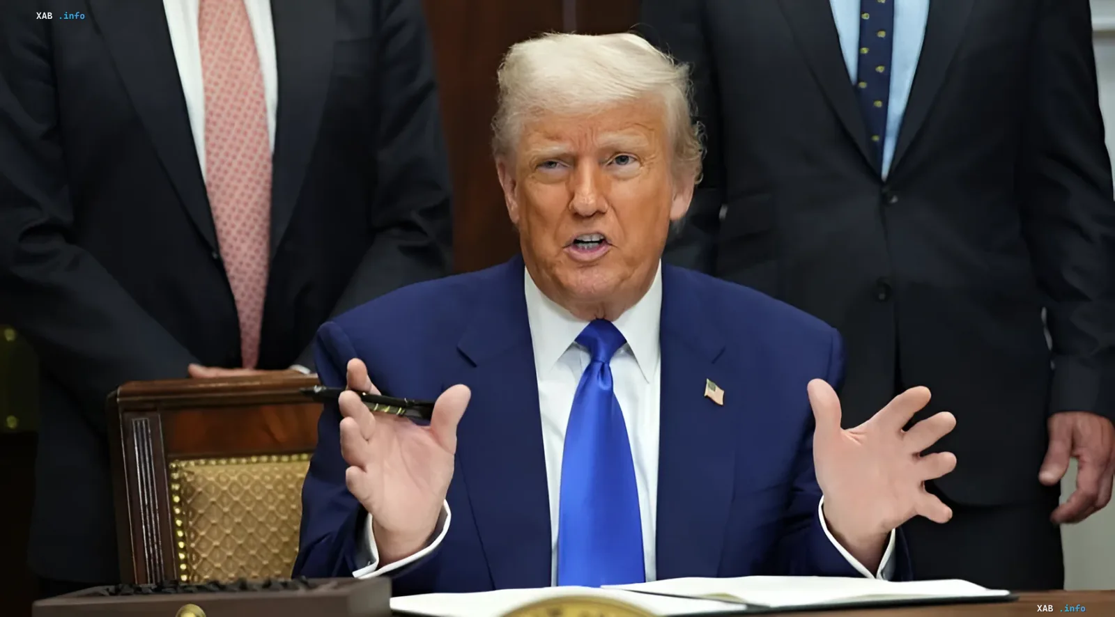 Donald Trump in a blue suit and tie, seated at a desk with documents, gesturing during a speech; two men in business suits stand behind him; visual for news on US pressure on Spain and EU resistance