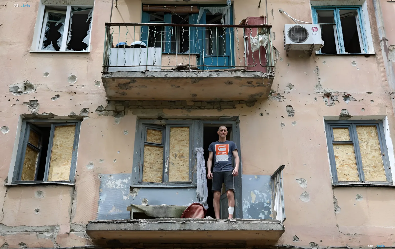A man stands on the balcony of a destroyed building with bullet holes in the walls and boarded-up windows, illustrating the aftermath of combat in Ukraine in June 2026.