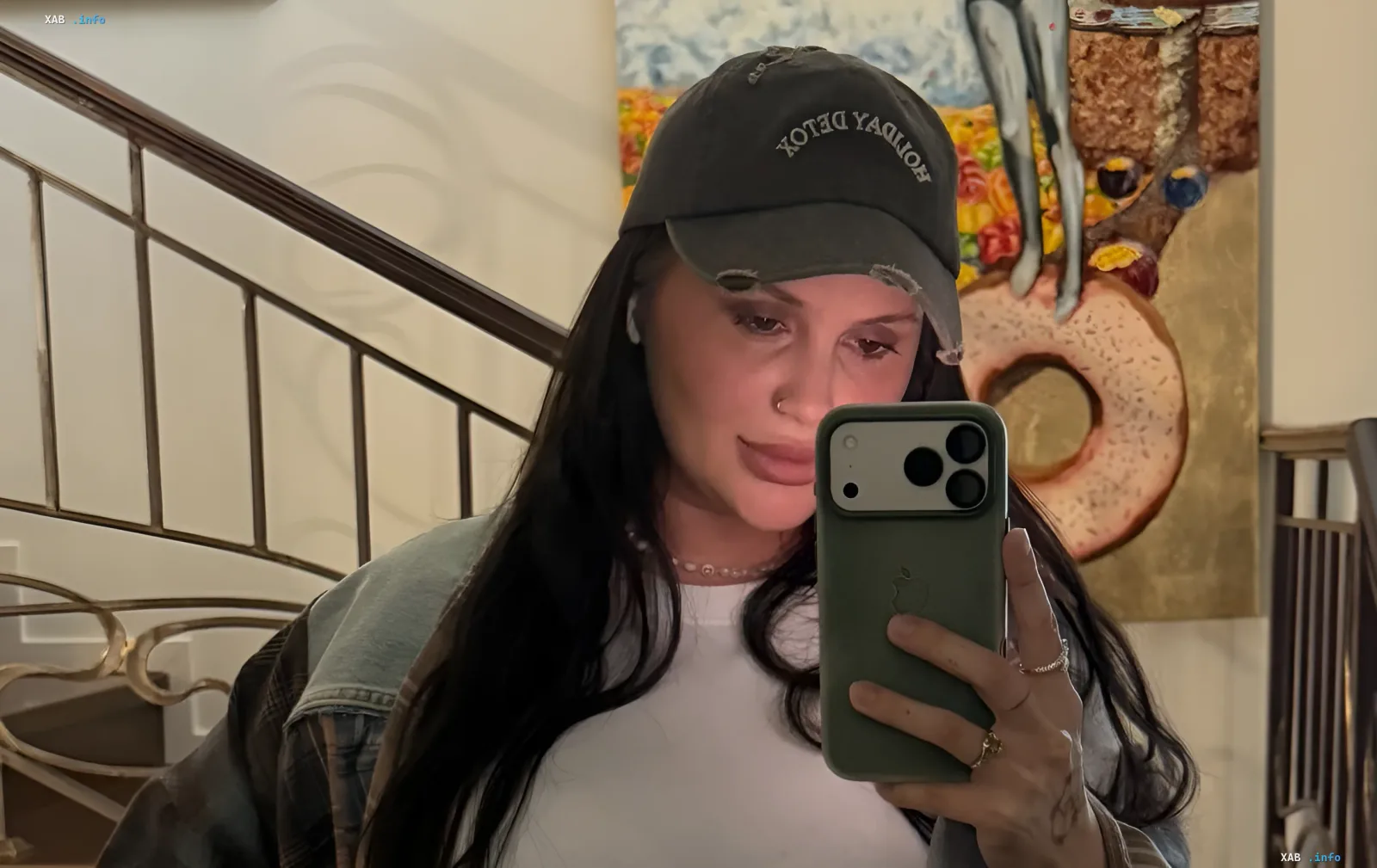 Алла Волкова в кепке с надписью 'HOLIDAY DETOX' делает селфи в зеркале, держа смартфон; на фоне — картина с абстрактным изображением