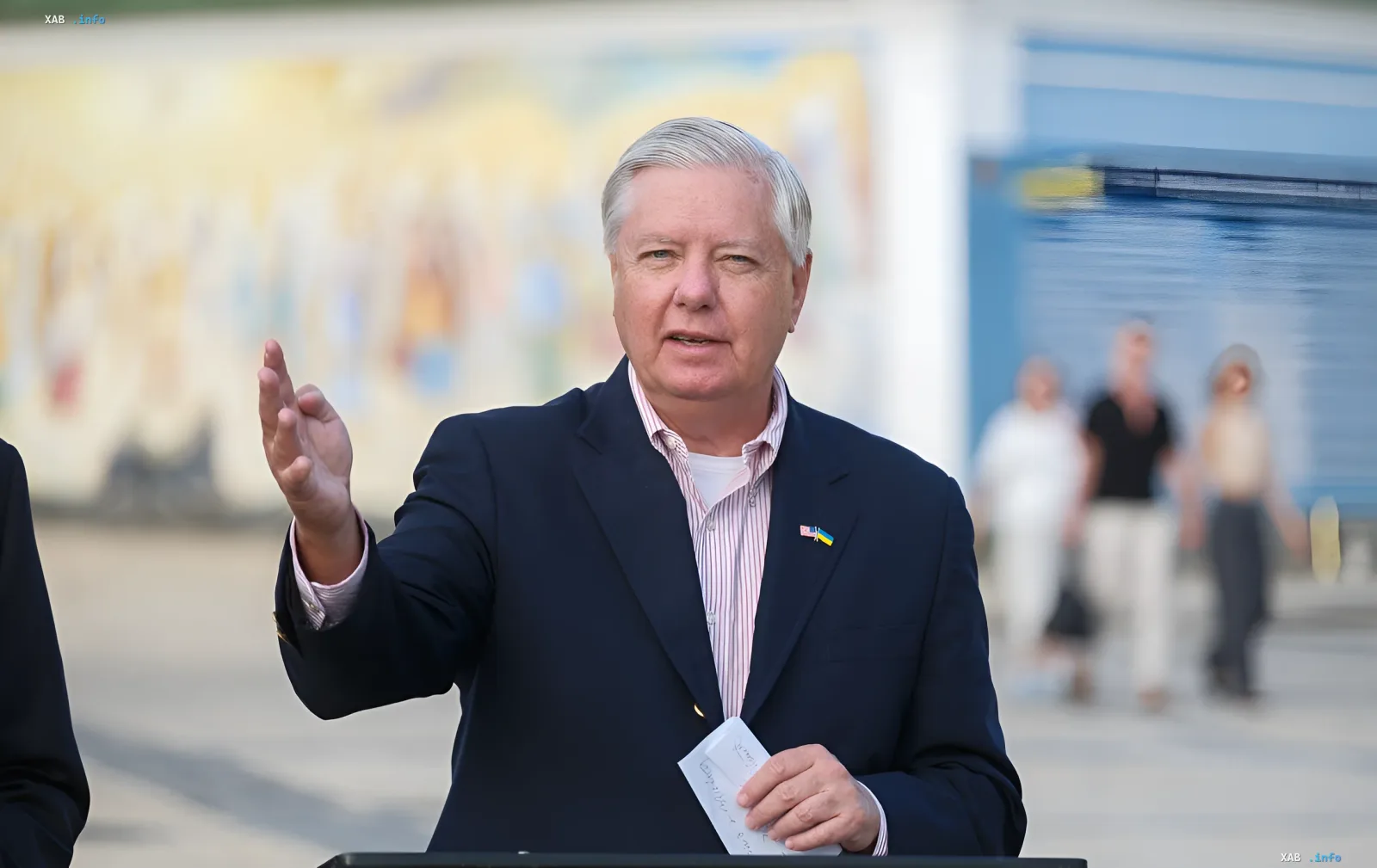 Сенатор Линдси Грэм выступает с речью об Украине, держа в руках заметки и жестикулируя, на фоне размытого городского пейзажа