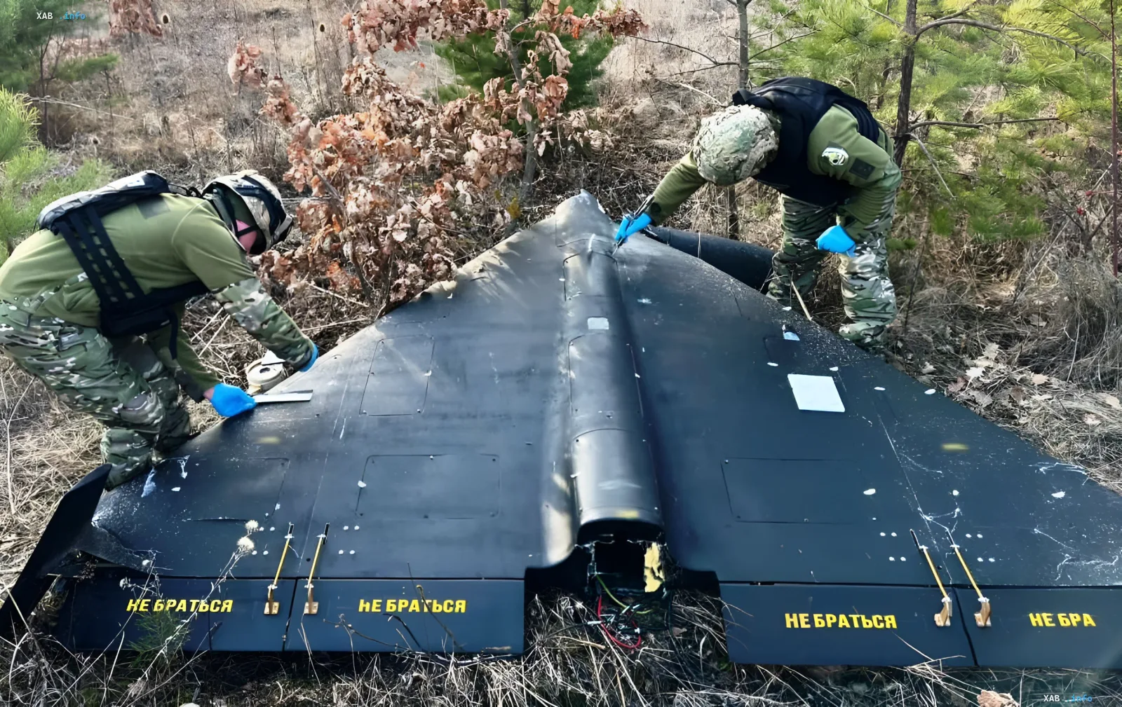 Российские военные осматривают обломки крупного черного дрона с надписями 'НЕ БРАТЬСЯ' в лесу, что подтверждает тему массового производства дронов для атак по логистике Украины.