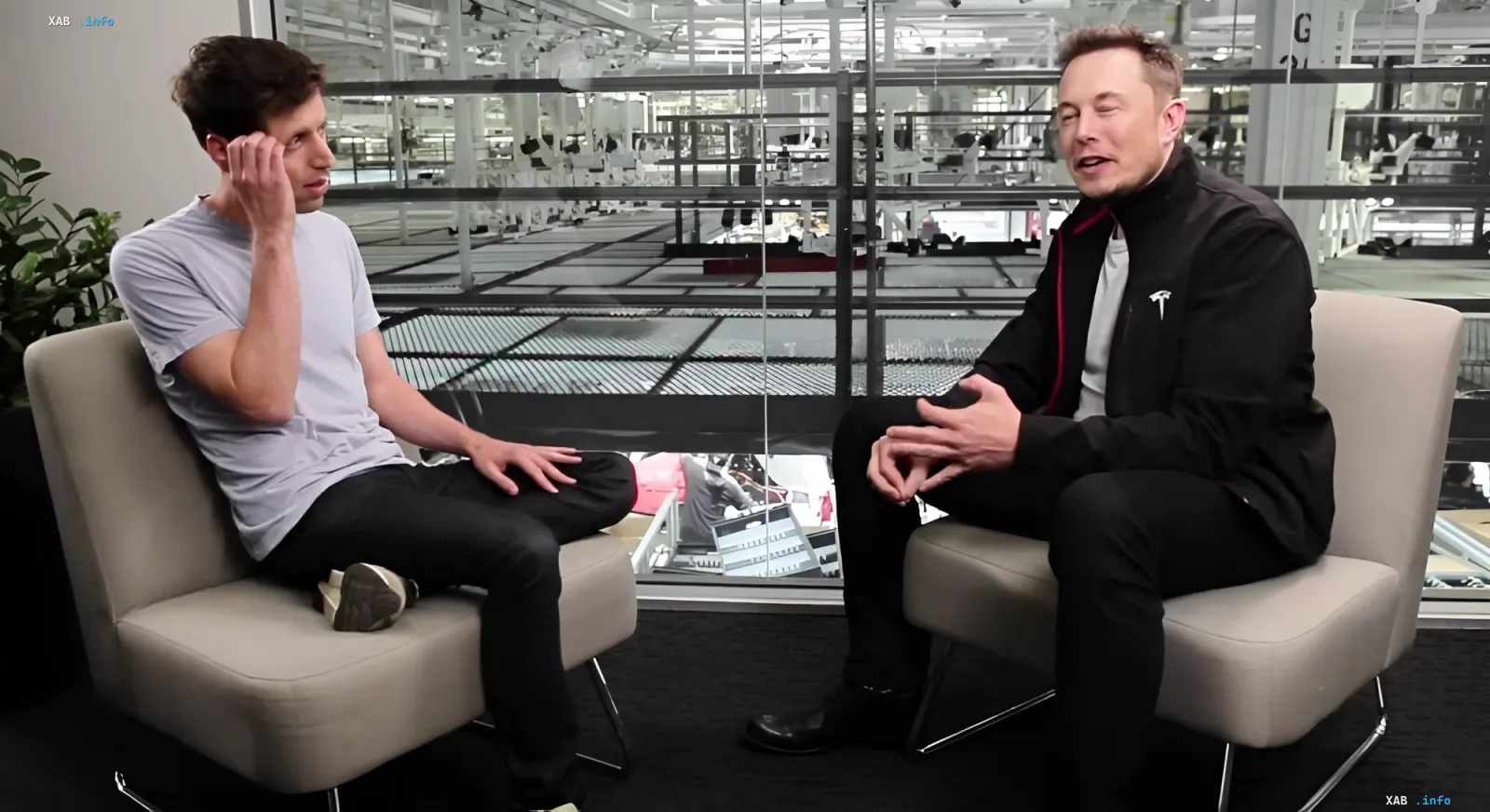 Elon Musk and Sam Altman sit in chairs in front of a glass wall overlooking a factory floor, discussing AI amid the Tesla-OpenAI conflict.