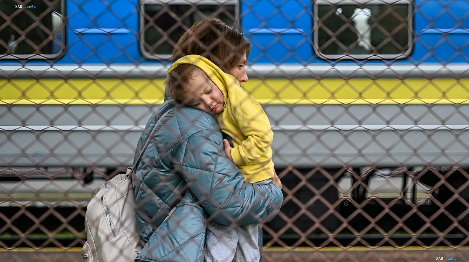 Mother holding a child in front of a train with Ukrainian colors behind a fence