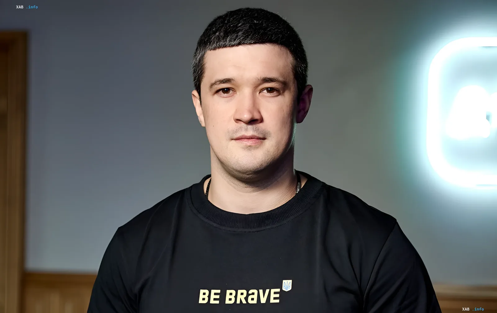 Michail Fedorow in einem schwarzen T-Shirt mit der Aufschrift 'BE BRAVE' und ukrainischem Wappen, vor einem leuchtenden Logo-Hintergrund