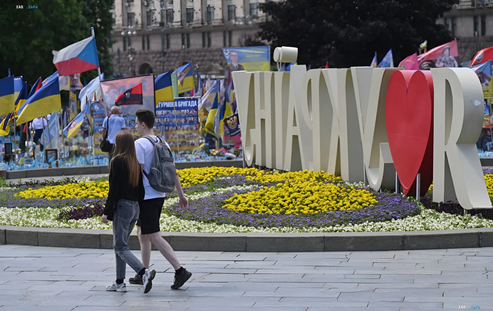 Молода пара гуляє по сонячному парку в Києві, поруч із квітником та пам'ятником «Я люблю Київ», на тлі українських прапорів — символ повернення спеки та літнього настрою в Україні