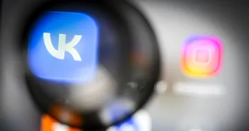 Логотип VK в увеличительном стекле на фоне размытого экрана смартфона с иконкой Instagram — иллюстрация к новости об удалении VK и Max из Google Play