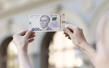 Жіночі руки тримають купюру номіналом 500 гривень на тлі розмитого міського пейзажу, ілюструючи тему парадоксу вартості гривні.