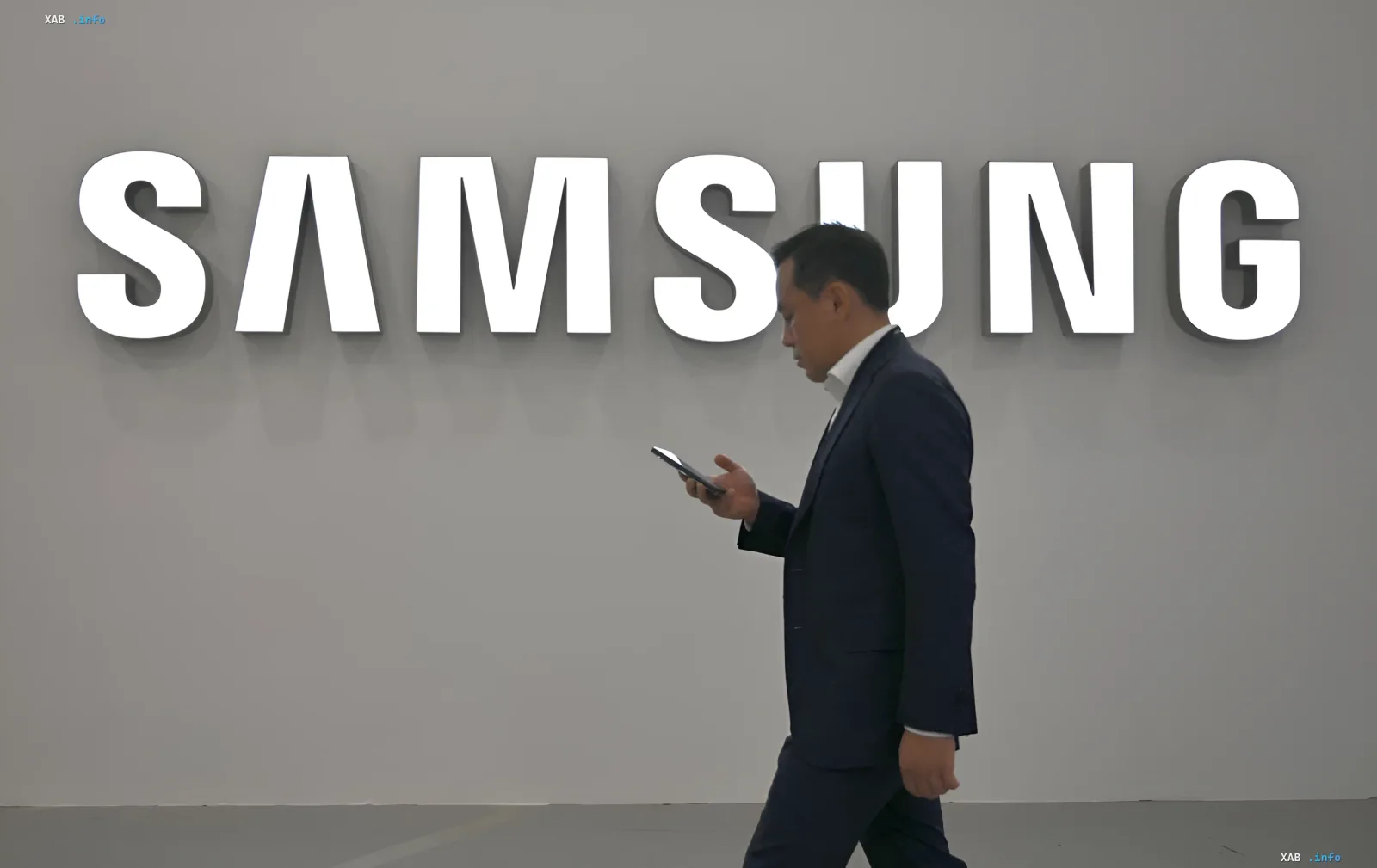 Мужчина в деловом костюме идет перед логотипом Samsung, держа смартфон в руке, иллюстрируя тему статьи о Samsung Galaxy A57 5G.