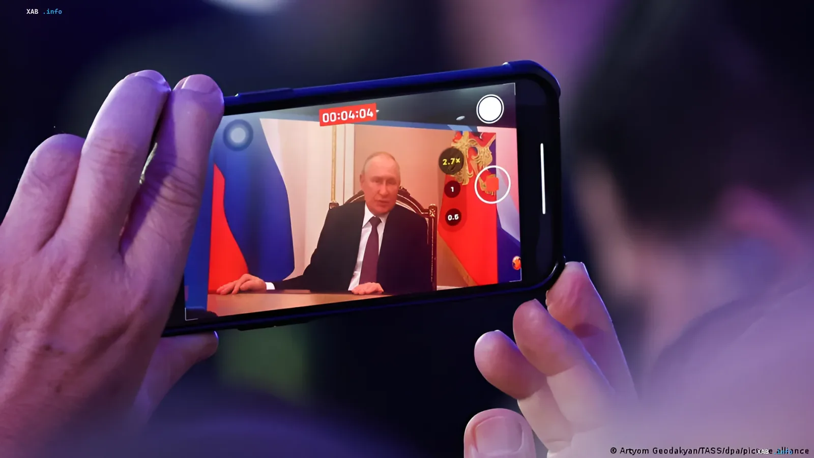 Человек фотографирует на смартфон выступление Владимира Путина, на экране видны флаги России, что иллюстрирует тему цифрового суверенитета и санкций ЕС против мессенджера «Макс»
