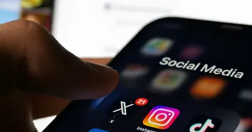Палец нажимает на папку «Social Media» на смартфоне, где видны иконки X, Instagram и TikTok — иллюстрация к новости о запрете соцсетей детям до 15 лет во Франции