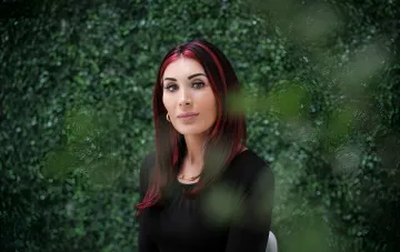 Laura Lumer mit roten Haaren vor einem Hintergrund aus grünem Laub, was ihre persönliche Erfahrung in der Ukraine symbolisiert