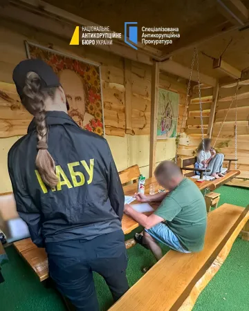 Співробітник НАБУ у формі з написом «НАБУ» на спині проводить допит або опитування в дерев'яному приміщенні, поруч сидить чоловік за столом з документами; логотипи НАБУ та САП зверху — ілюстрація до розслідування схеми на 150 млн гривень у Нацгвардії