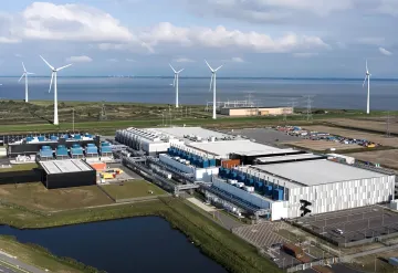 Aerial view of a modern data center with wind turbines in the background, symbolizing the massive energy consumption and hidden financial obligations of AI giants.