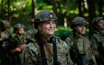 Female Ukrainian soldier in camouflage uniform and helmet with new body armor, standing in forest among comrades — first batch of specialized body armor for women has entered service