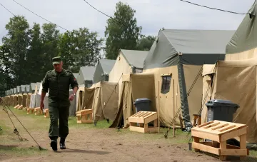Военнослужащий в форме проходит мимо рядов палаток в военном лагере, иллюстрируя подготовку к мобилизации на жатву в Беларуси.