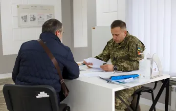 Український військовий у камуфляжному одязі переглядає документи за столом, поки цивільний чоловік у синій куртці чекає рішення — ілюстрація до теми скасування Координаційного центру з бронювання