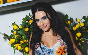 Анна Тринчер в ярком наряде с цветочными узорами на фоне лимонных ветвей — центральная фигура скандала вокруг песни «Маргарита»