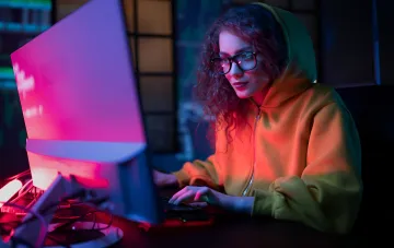 Hacker in einer Kapuze arbeitet an einem Computer in einem dunklen Raum mit Neonbeleuchtung, was einen Cyberangriff auf den Bildungssektor symbolisiert