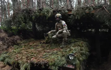 Российский военнослужащий в камуфляже и защитном шлеме, маскирующийся под еловыми ветками в лесу Харьковской области