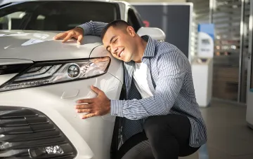 Glücklicher Mann umarmt ein weißes Auto im Showroom, was die wachsende Nachfrage nach Benzin- und Hybridfahrzeugen in der Ukraine symbolisiert