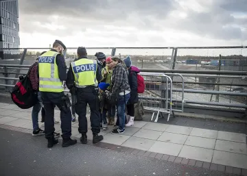 Schwedische Polizisten in Westen mit der Aufschrift POLIS im Gespräch mit einer Gruppe von Migranten auf einer Brücke, was die Verschärfung der Migrationskontrolle veranschaulicht
