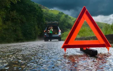 Frau mit Kind neben defektem Auto auf der Straße, Warnzeichen in der Nähe, dunkles Wetter