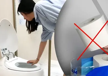 Frau untersucht eine intelligente Toilette mit Bidet-Funktion, während eine Toilettenpapierrolle mit rotem Kreuz durchgestrichen ist — Symbol für das Ende der Papierära