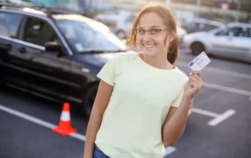 Happy Ukrainian woman with glasses holding a driver’s license against a parking lot backdrop, symbolizing successful acquisition of driving rights without retaking exams