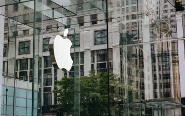 Apple-Logo auf einer Glasfassade eines Geschäfts, das städtische Gebäude widerspiegelt und Innovation sowie Datenschutz bei der Entwicklung intelligenter Brillen symbolisiert.