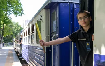 Юный машинист в форме Укрзализныцы держит желтый флажок у двери поезда на детской железной дороге Украины
