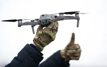 Military personnel in camouflage gloves holding a drone, symbolizing the US lag in drone production compared to Ukraine