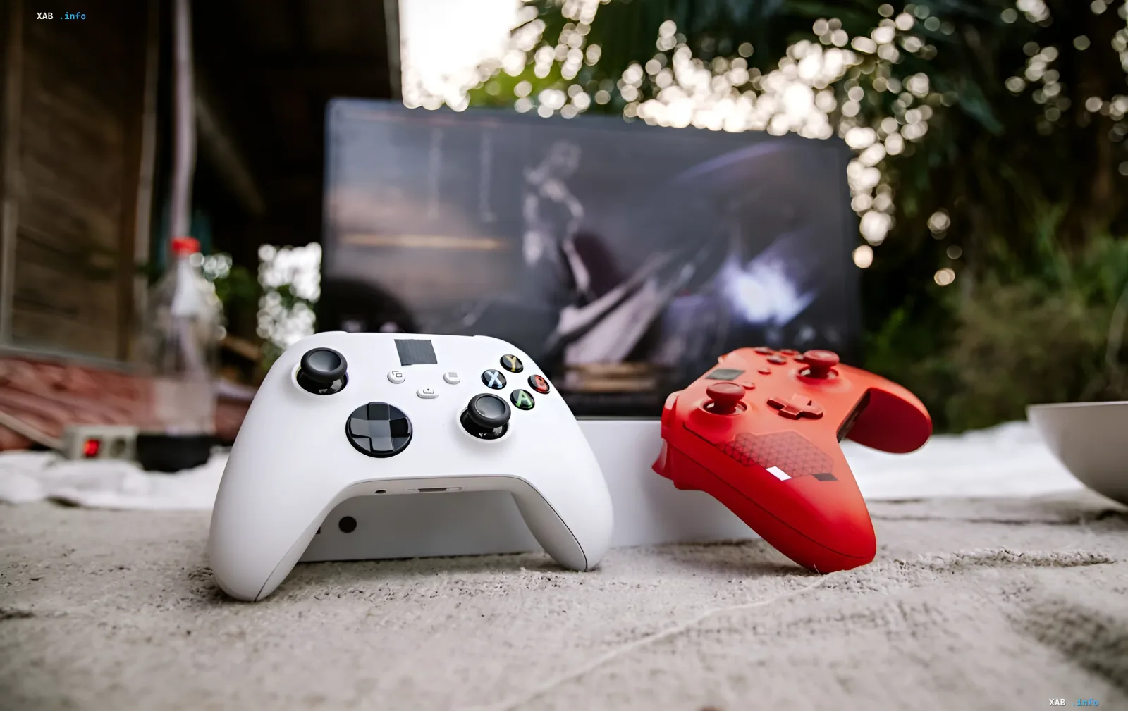 Белый и красный геймпады Xbox на переднем плане с размытым экраном игры на заднем фоне, иллюстрирующие доступность игр Xbox на ПК благодаря партнерству с Ubisoft