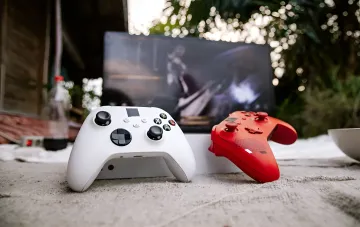 Белый и красный геймпады Xbox на переднем плане с размытым экраном игры на заднем фоне, иллюстрирующие доступность игр Xbox на ПК благодаря партнерству с Ubisoft