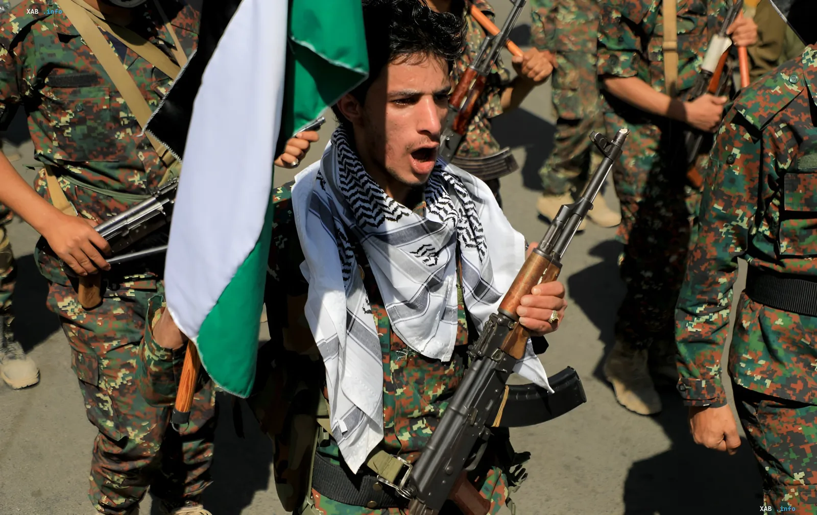 Houthi fighter with an assault rifle and Yemeni flag in front of a group of armed fighters in camouflage