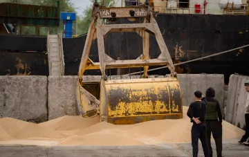 Bagger lädt Getreide in einem Hafen mit einem Frachtschiff im Hintergrund, was die Bedrohung des Exports durch Angriffe auf ukrainische Häfen veranschaulicht.