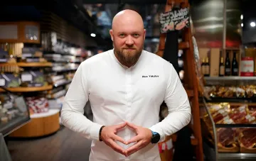 Алекс Якутов в белом кителе с вышитым именем, стоящий в гастрономическом магазине и делающий жест сердца руками — символ любви к сыну после усыновления